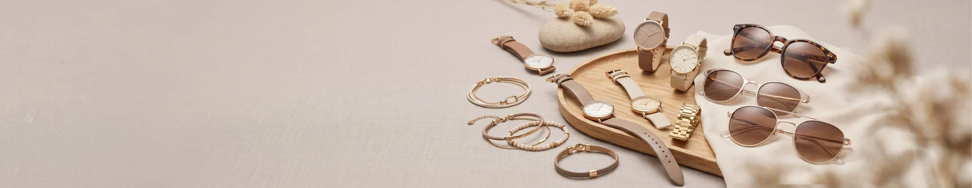 A styled flat-lay of various fashion accessories on a light beige background, including multiple pairs of sunglasses, several gold-tone watches, bracelets, rings, and a wooden tray.