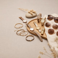 A styled flat-lay of multiple fashion accessories on a neutral beige fabric background, including several wristwatches with leather and metal straps, multiple pairs of sunglasses with brown and tortoiseshell lenses, several gold-tone bracelets, and a small wooden tray with decorative stones and dried grass.