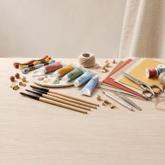A styled flat-lay of craft supplies on a light wood table, including paintbrushes, tubes of acrylic paint, thread, twine, scissors, tweezers, wooden beads, and colored paper sheets.