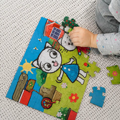 A child's hand is assembling a colorful cartoon jigsaw puzzle featuring a white cat and a blue dress, placed on a textured white woven blanket.