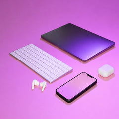 A styled flat-lay of electronic accessories on a pink and purple gradient background, including a closed laptop, a white wireless keyboard, white earbuds, a smartphone, and a white charging case.