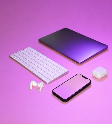 A styled flat-lay of electronic accessories on a pink and purple gradient background, including a closed laptop, a white wireless keyboard, white earbuds, a smartphone, and a white charging case.