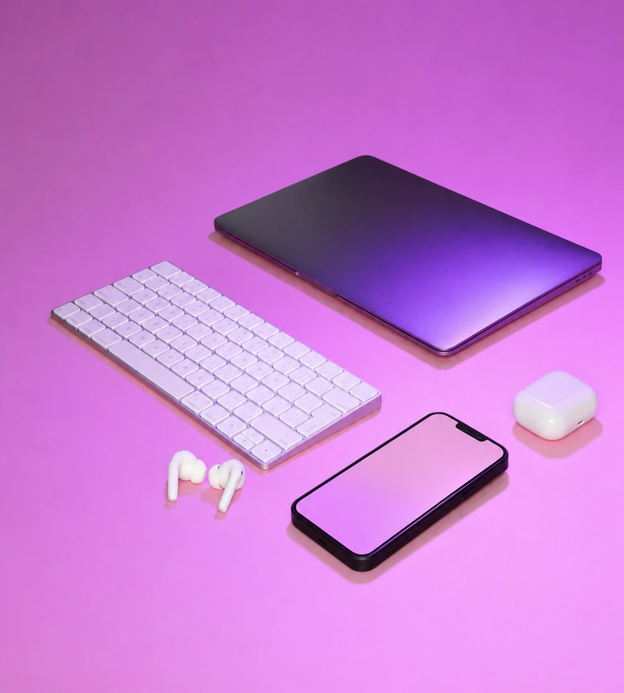 A styled flat-lay of electronic accessories on a pink and purple gradient background, including a closed laptop, a white wireless keyboard, white earbuds, a smartphone, and a white charging case.