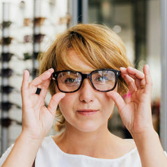 A woman with short blonde hair holds a pair of black rectangular eyeglasses up to her eyes in what appears to be an optician's shop.