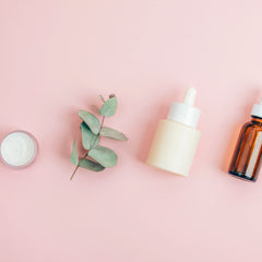 A styled flat-lay of skincare products on a solid pink background, including a frosted cream bottle with a white dropper, an amber glass serum bottle, a small white jar, and a green eucalyptus sprig.