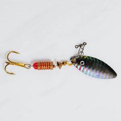 A fishing lure with two gold treble hooks, a red bead, and a metallic spinnerbait body with an iridescent blue, green, and silver finish, photographed on a white marble background.