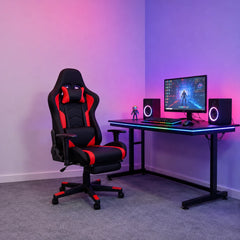 A black and red gaming chair positioned next to a black desk setup with a monitor, keyboard, speakers, and ambient pink and purple LED lighting in a room corner.