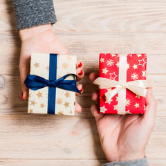 Two hands holding small wrapped gift boxes over a light wood tabletop. The left box is cream with gold stars and a dark blue ribbon, and the right box is red with white stars and a cream ribbon.