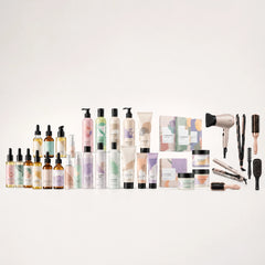 A collection of haircare products and styling tools arranged in rows on a clean white background. The set includes various bottles, tubes, jars, and boxes with pastel-colored labels, alongside a hair dryer, hairbrush, curling iron, and other accessories.