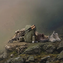 A hiking setup arranged on a rocky cliffside in a misty outdoor environment, including an olive green backpack, trekking poles, a water bottle, a sleeping pad, hiking boots, and a pair of trail shoes.