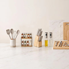 A styled kitchen countertop scene with wooden and stainless steel cookware and accessories, including a utensil holder, spice rack, knife block, oil and vinegar bottles, and cutting boards, set against a warm off-white wall and light marble surface.