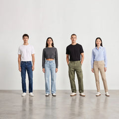 Four models standing in a row against a minimalist white studio wall and gray floor, wearing different casual and work-style outfits including jeans, cargo pants, a short-sleeve crop top, a long-sleeve button-up shirt, and chinos.