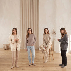 Four women standing in a minimalist room with beige walls and curtains, wearing coordinated neutral-toned loungewear and cardigans. The outfits include cream, taupe, beige, gray, and black tones, with one seated on a wooden stool and one holding a small cup.