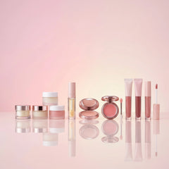 A collection of pink and rose gold cosmetic products, including jars, tubes, and compacts, arranged in a row on a reflective pink surface against a soft pink background.