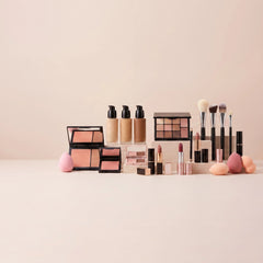 A cosmetics set arranged on a clean pale pink surface against a matching soft pink background. The set includes several foundation and concealer bottles, two open face palettes with peach and pink shades, a multi-shade eyeshadow palette, makeup brushes with black and white handles, and several pink makeup sponges.