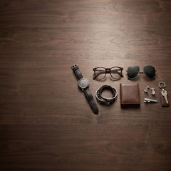 A styled flat-lay of men's accessories arranged on a dark brown wood-grain surface, including a black leather wristwatch, eyeglasses, sunglasses, a beaded bracelet, a brown leather wallet, earrings, a key, and a keychain.