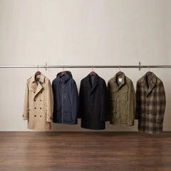 Five men's overcoats in beige, navy, black, olive green, and brown plaid hang on a metal rail against a light beige wall and dark wood floor.