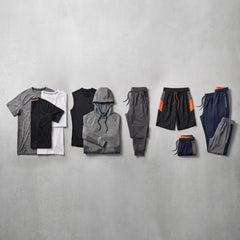 A flat-lay arrangement of athletic apparel on a light gray textured wall, including layered t-shirts, a sleeveless top, a hoodie, joggers, shorts, and sweatpants in gray, black, navy, and white with small orange accents.