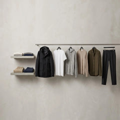 A minimalist clothing display featuring a black jacket, white t-shirt, gray hoodie, beige sweater, olive shirt, and dark trousers hanging on a rail, alongside floating shelves with folded garments, set against a textured light gray wall.