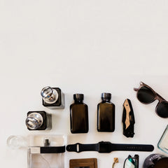 A styled flat-lay of various everyday carry accessories on a white surface, including two dark amber glass bottles, a folding pocket knife, sunglasses, a smartwatch band, a wallet, and small bottles.
