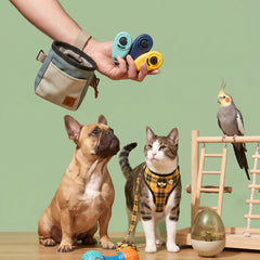 A lifestyle scene featuring a French Bulldog and a tabby cat wearing harnesses, with a human hand holding two pet cameras and a small pouch. A cockatiel is perched on a wooden toy ladder against a solid sage green background.