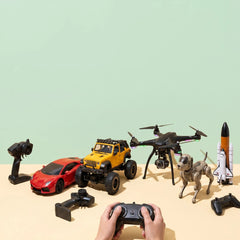 A collection of remote control toys arranged on a beige surface against a pale green background, including a red sports car, yellow off-road truck, black quadcopter drone, robotic dog, space shuttle model, and several controllers. A pair of hands holds a black controller in the foreground.