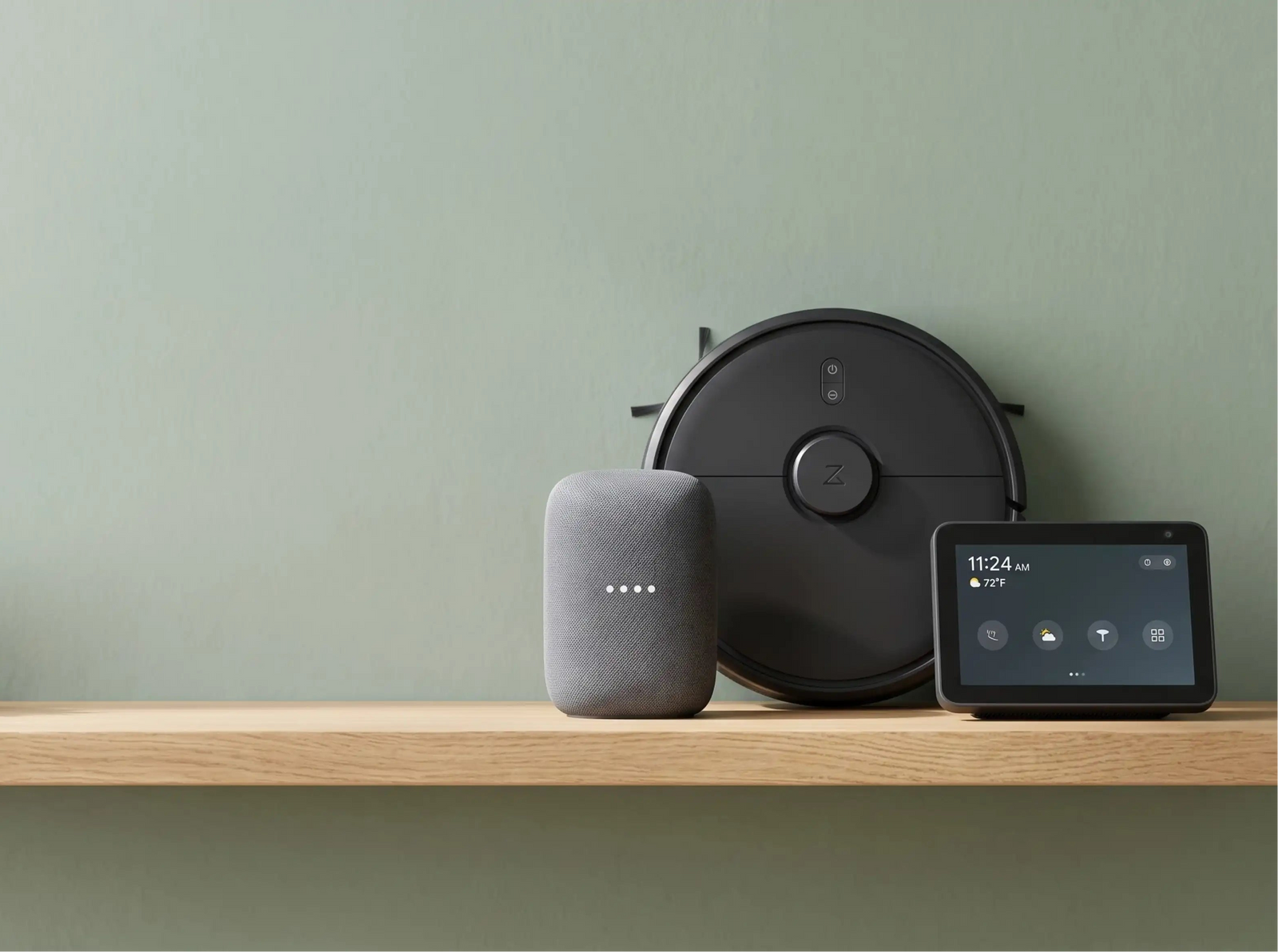 A smart home setup on a light wood shelf against a sage green wall, featuring a black robotic vacuum, a gray smart speaker, and a black tablet display, with a small potted succulent on the left.