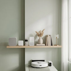 A modern home interior scene with a light wood floating shelf mounted on a sage green wall. The shelf holds several minimalist white and beige home electronics and decor items, including a small vase with dried grass, books, and a lamp.