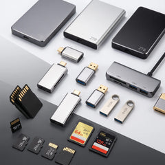 A collection of portable electronic accessories arranged on a split light and dark gray background, including multiple USB flash drives, SD memory cards, and external storage devices in silver, black, and gold finishes.