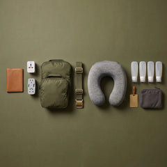 A travel packing set arranged neatly on a solid olive green background, including an olive green backpack, a gray U-shaped neck pillow, a brown leather notebook, white adapters, plastic bottles, a gray pouch, and a brown luggage tag.