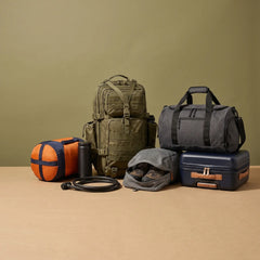 A collection of outdoor and travel gear arranged on a beige surface against a muted olive-green background, including a large olive green backpack, a dark gray duffel bag, a navy hard case, an orange sleeping bag, a gray pouch with shoes, and a black bottle and hose.