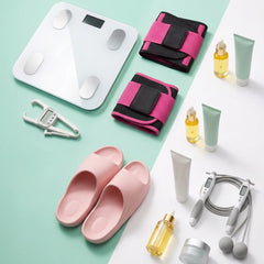 A styled flat-lay of fitness and wellness items on a split mint green and white background, including a white digital body scale, two pink-and-black wrist wraps, a silver hook, a pair of light pink slip-on slippers, a jump rope, small bottles and tubes, and a small jar.