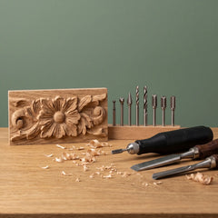 Woodworking tools on a wooden table including a rotary chisel, two hand chisels, a drill bit set in a wooden holder, and a carved wooden decorative block with a floral relief, set against a muted green background.