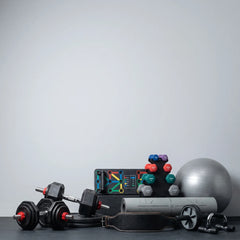 A collection of fitness equipment arranged on a dark floor against a light gray wall, including adjustable dumbbells, a weight bench, a foam roller, a black wrist strap, a silver stability ball, and a stack of colorful dumbbells.
