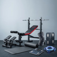A fitness equipment set arranged on a dark floor against a light gray wall, including a weight bench with red padding, a barbell rack, a knee support, a resistance band, a foam roller, a mat, and a digital scale.