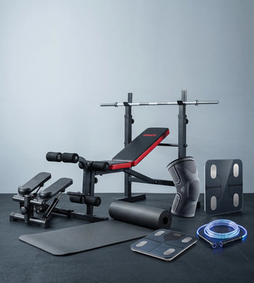 A fitness equipment set arranged on a dark floor against a light gray wall, including a weight bench with red padding, a barbell rack, a knee support, a resistance band, a foam roller, a mat, and a digital scale.