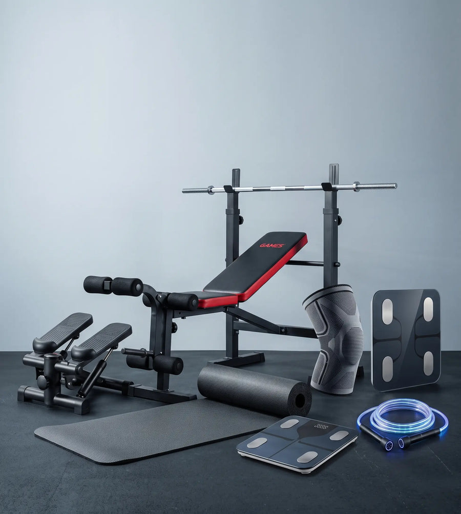 A fitness equipment set arranged on a dark floor against a light gray wall, including a weight bench with red padding, a barbell rack, a knee support, a resistance band, a foam roller, a mat, and a digital scale.