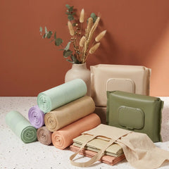 A styled product display of reusable baby care items including rolled diapers, a diaper bag, wipes, and a diaper pail cover, arranged on a speckled white surface against a terracotta background with a vase of dried grasses.